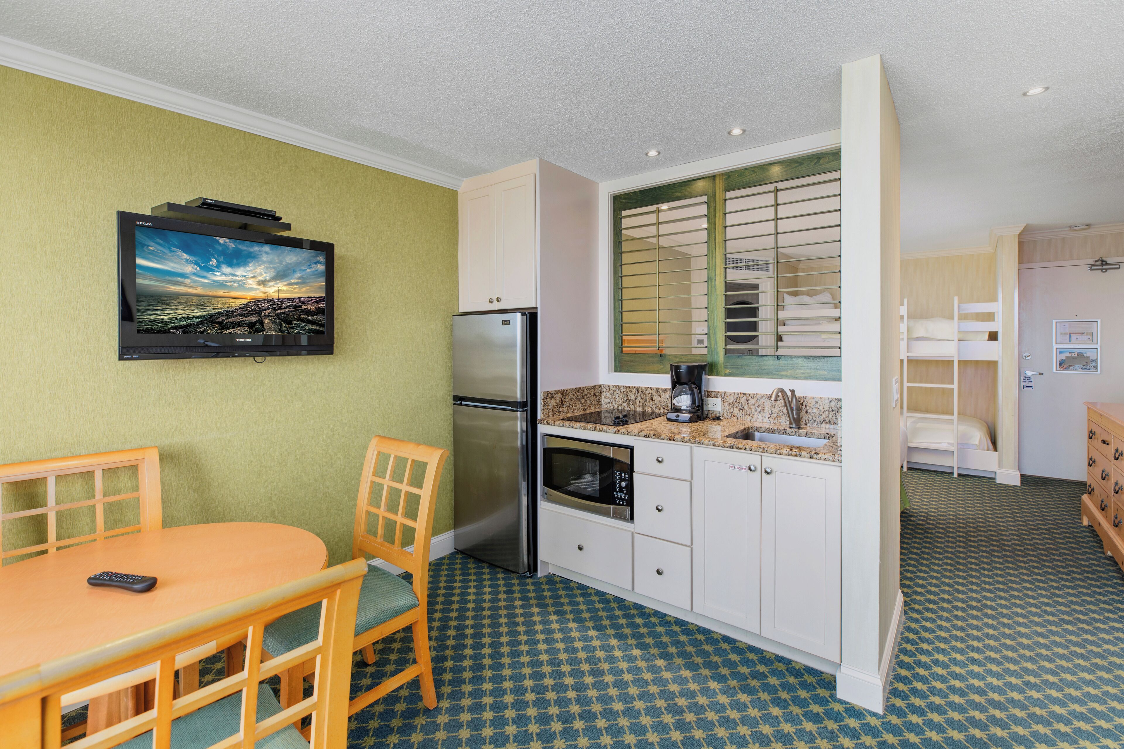 A compact kitchen with a refrigerator, microwave, and a small countertop. There is a dining table with chairs and a television mounted on the wall.