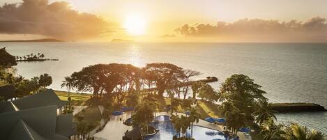 A resort pool area with palm trees and umbrellas, overlooking the sea at sunset.