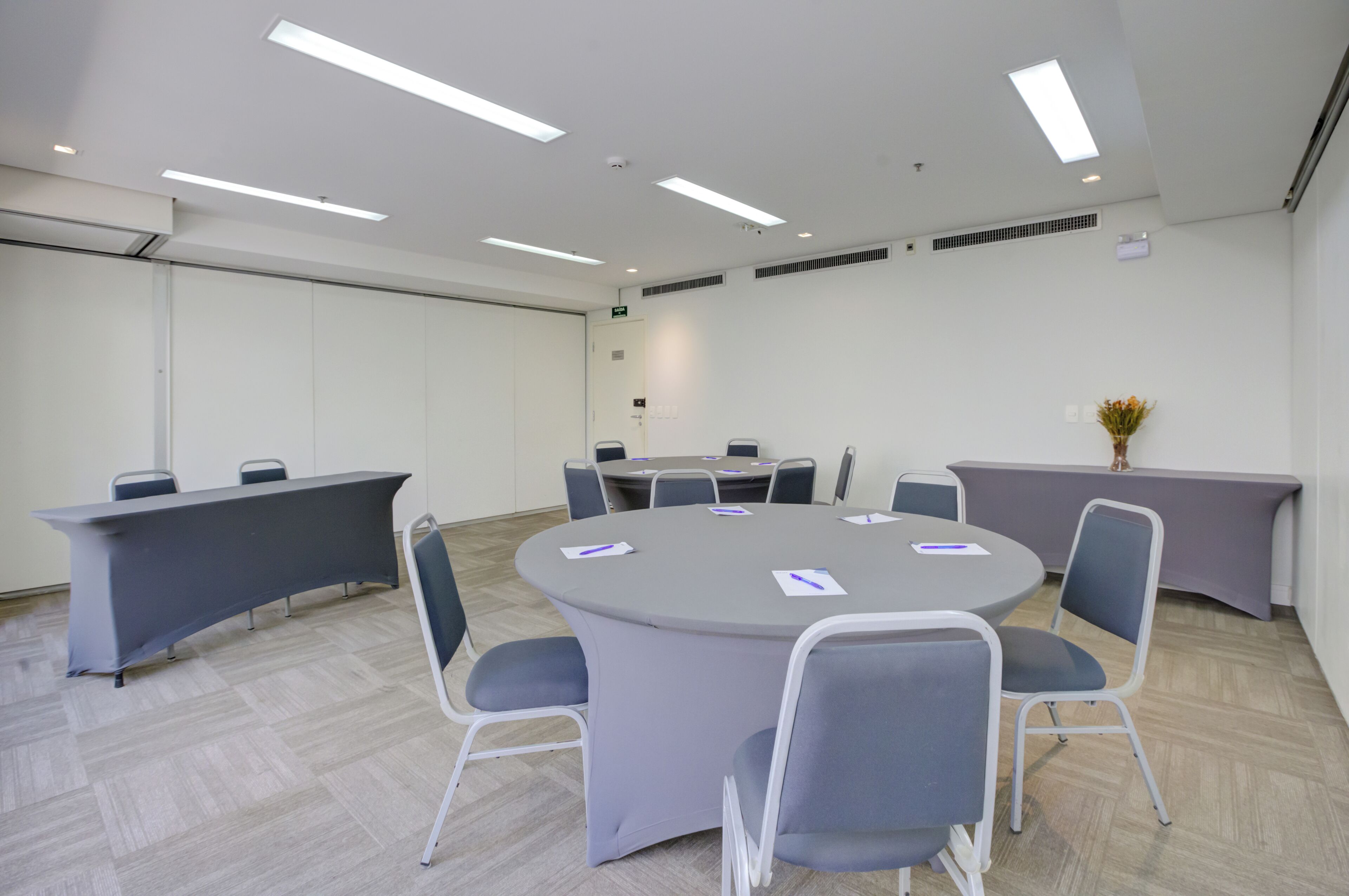 A conference room with a round table, grey chairs, a long table with a grey tablecloth, and a vase with flowers on the wall.