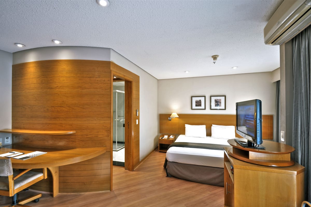 Habitación de hotel con escritorio de madera, cama, televisión y zona de ducha.