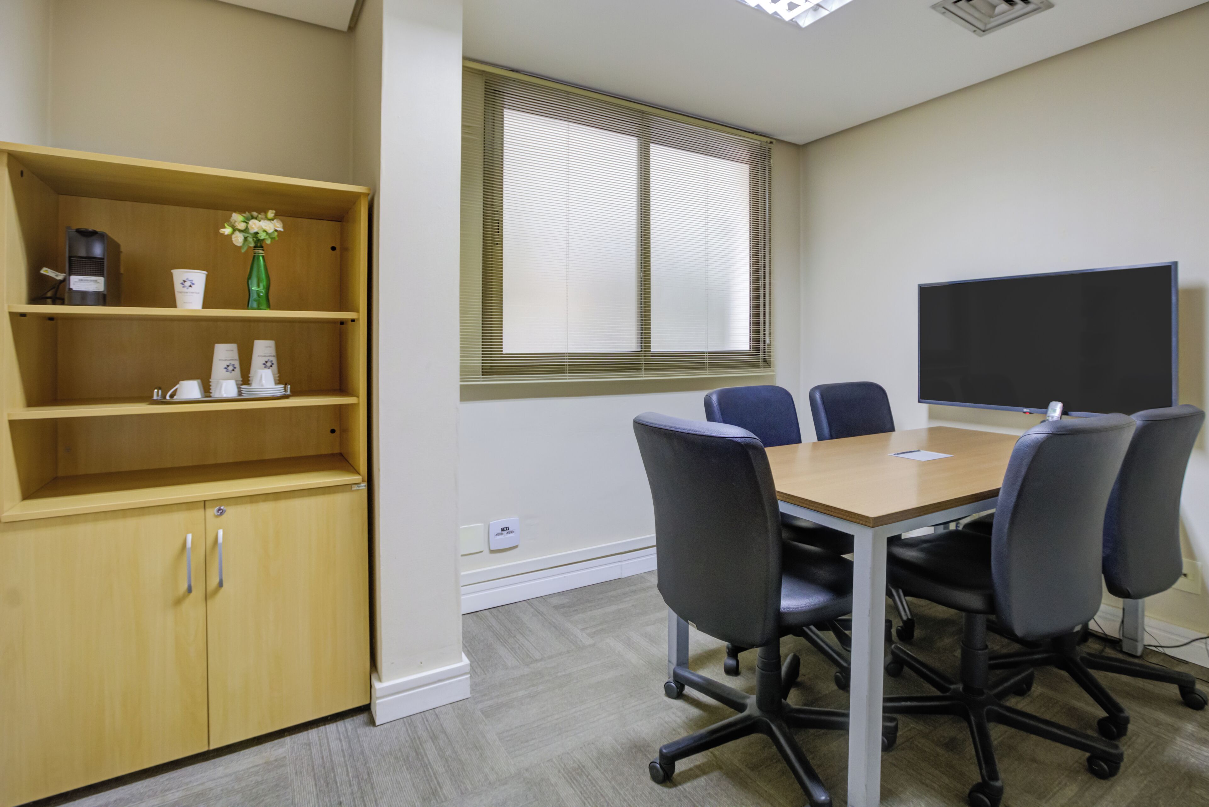 Un salón de conferencias con una mesa de madera, cuatro sillas, un televisor de pantalla plana grande, una estantería con tazas de café y un jarrón, y una ventana con persianas.