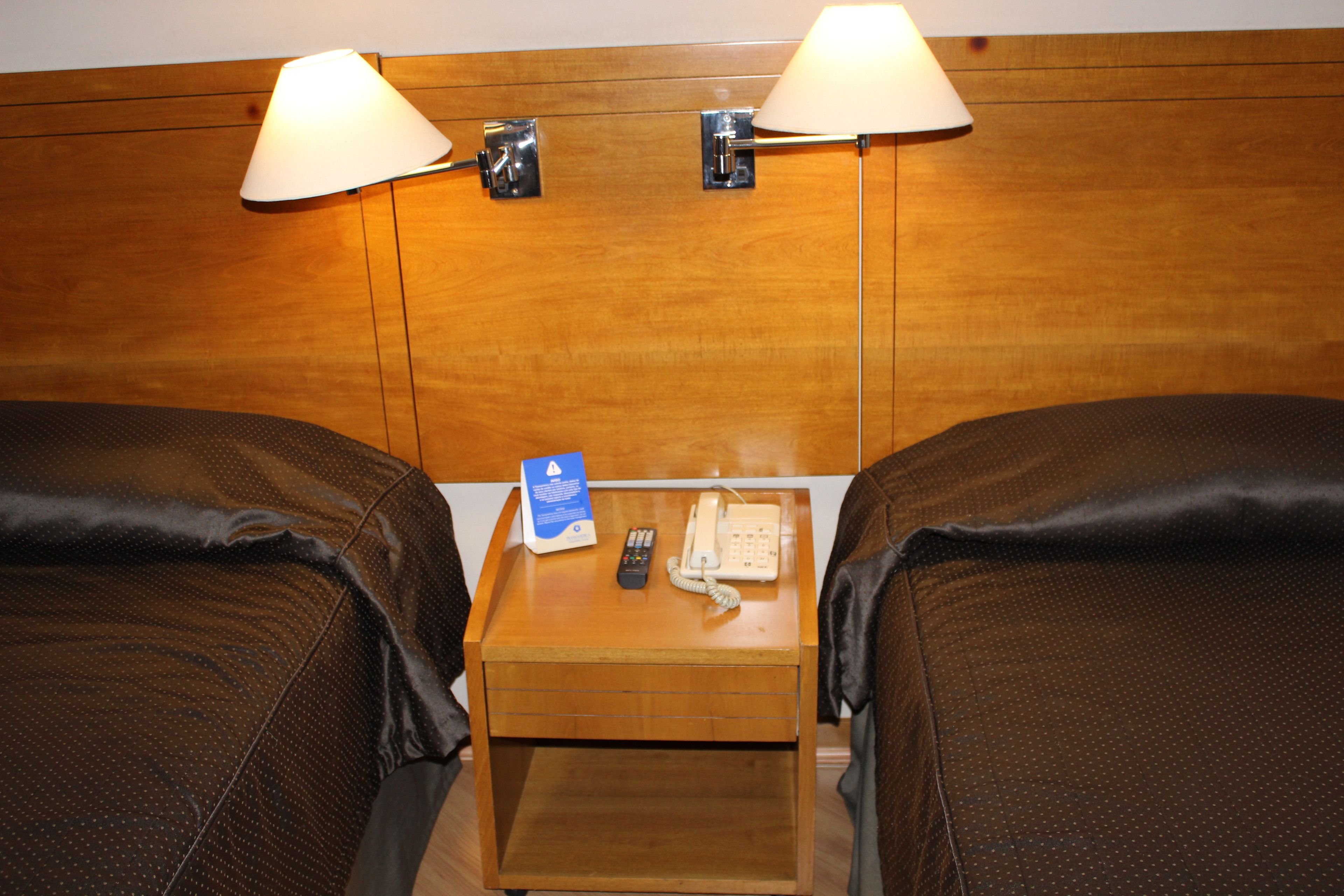 Ein Hotelzimmer mit zwei Betten, einem hölzernen Kopfteil, Nachttischlampen, einem Nachttisch mit Telefon und Fernbedienung sowie einer blauen Informationstafel.