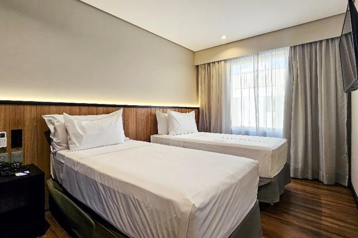 A hotel room with two beds, a TV mounted on the wall, and a window with curtains.