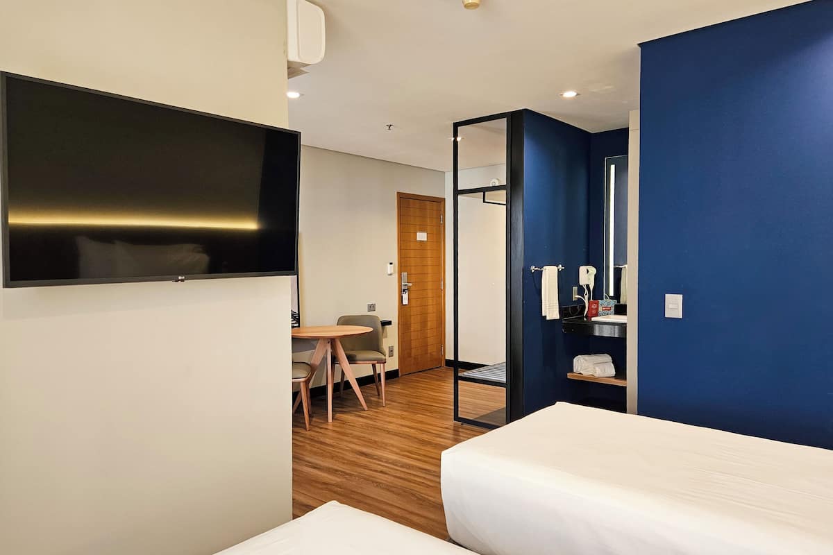 Una habitación de hotel con cama, escritorio con silla, televisor montado en la pared y espejo.