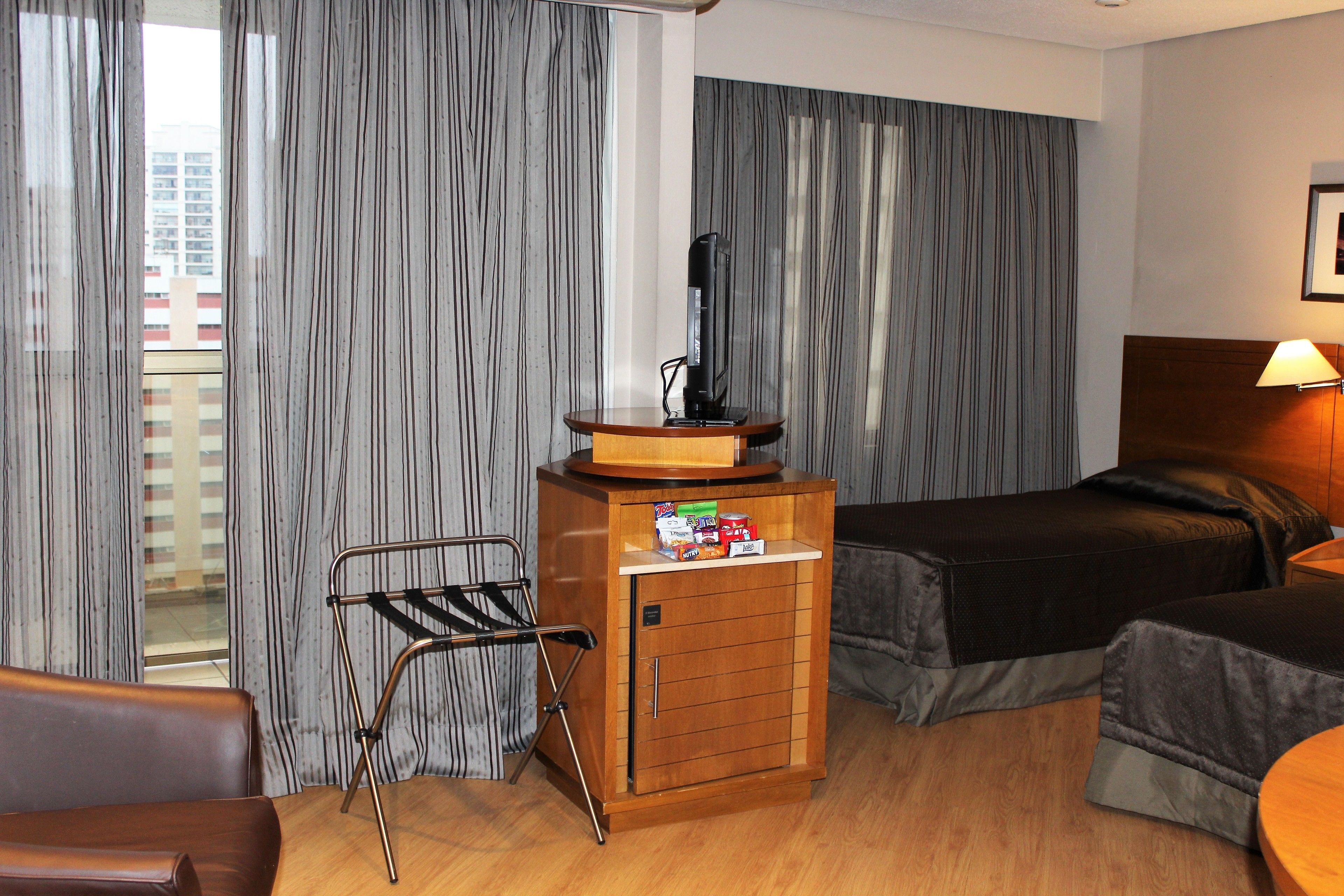 Una camera d'albergo con un letto, un piccolo mobiletti in legno con articoli da toilette, una sedia pieghevole e una finestra con tende.