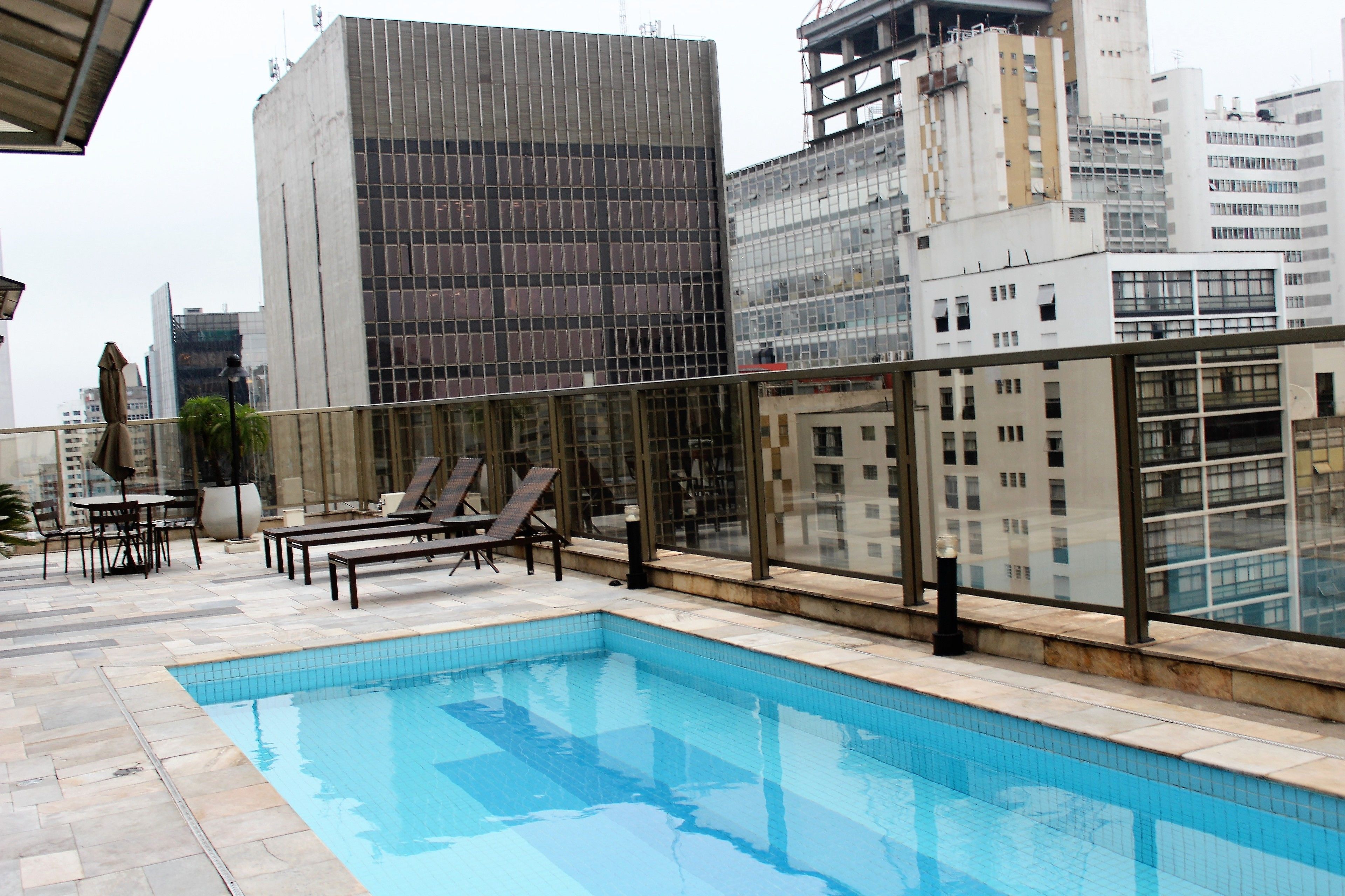 Una piscina en la azotea con sillas de descanso y barandilla de cristal, rodeada de edificios altos.