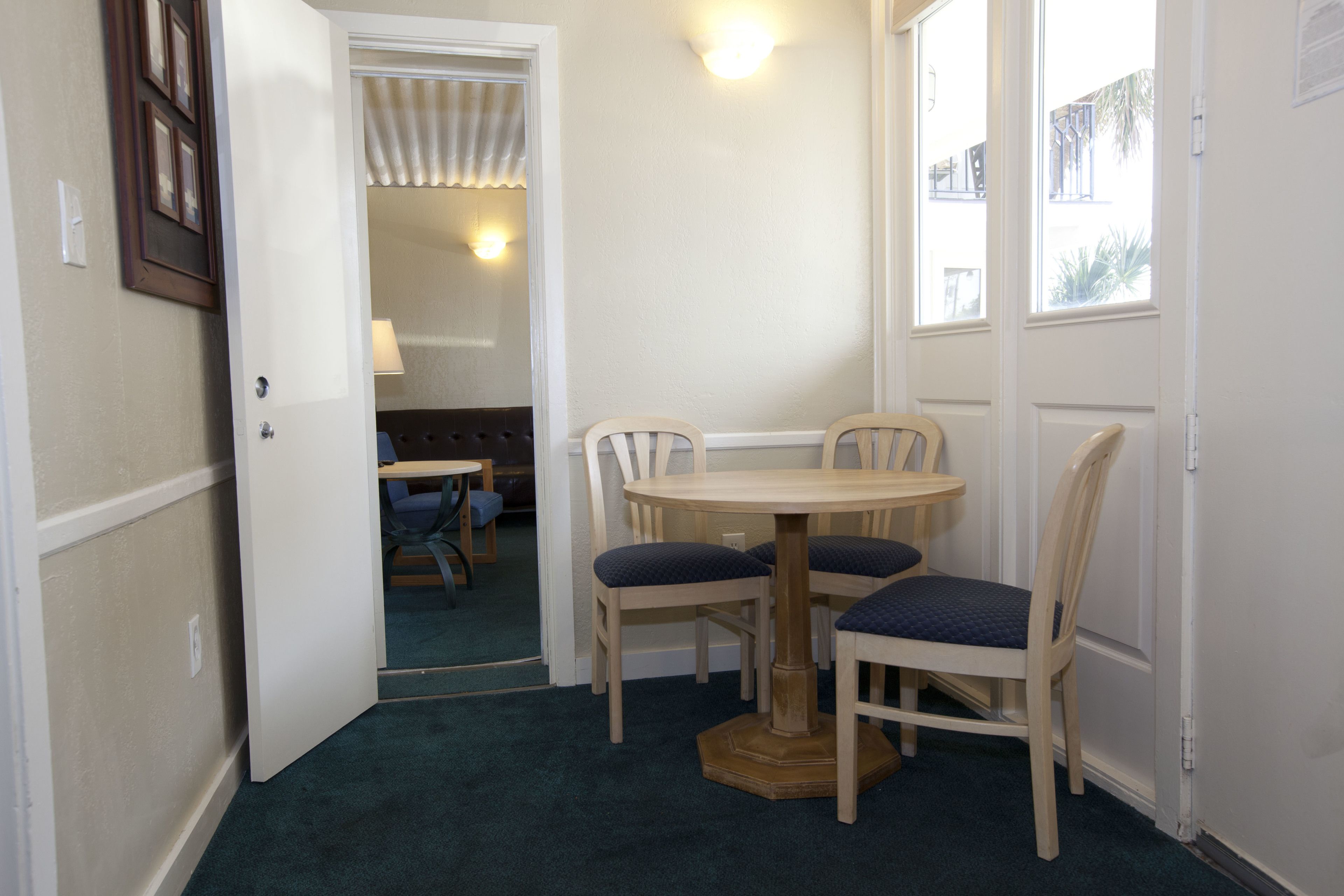 Um quarto pequeno com uma mesa redonda e quatro cadeiras, uma porta que dá para outro espaço, e uma janela com vista para edifícios.