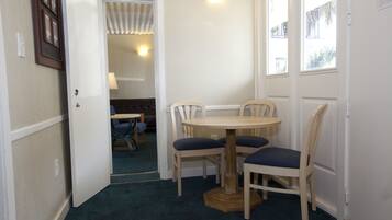 Um quarto pequeno com uma mesa redonda e quatro cadeiras, uma porta que dá para outro espaço, e uma janela com vista para edifícios.