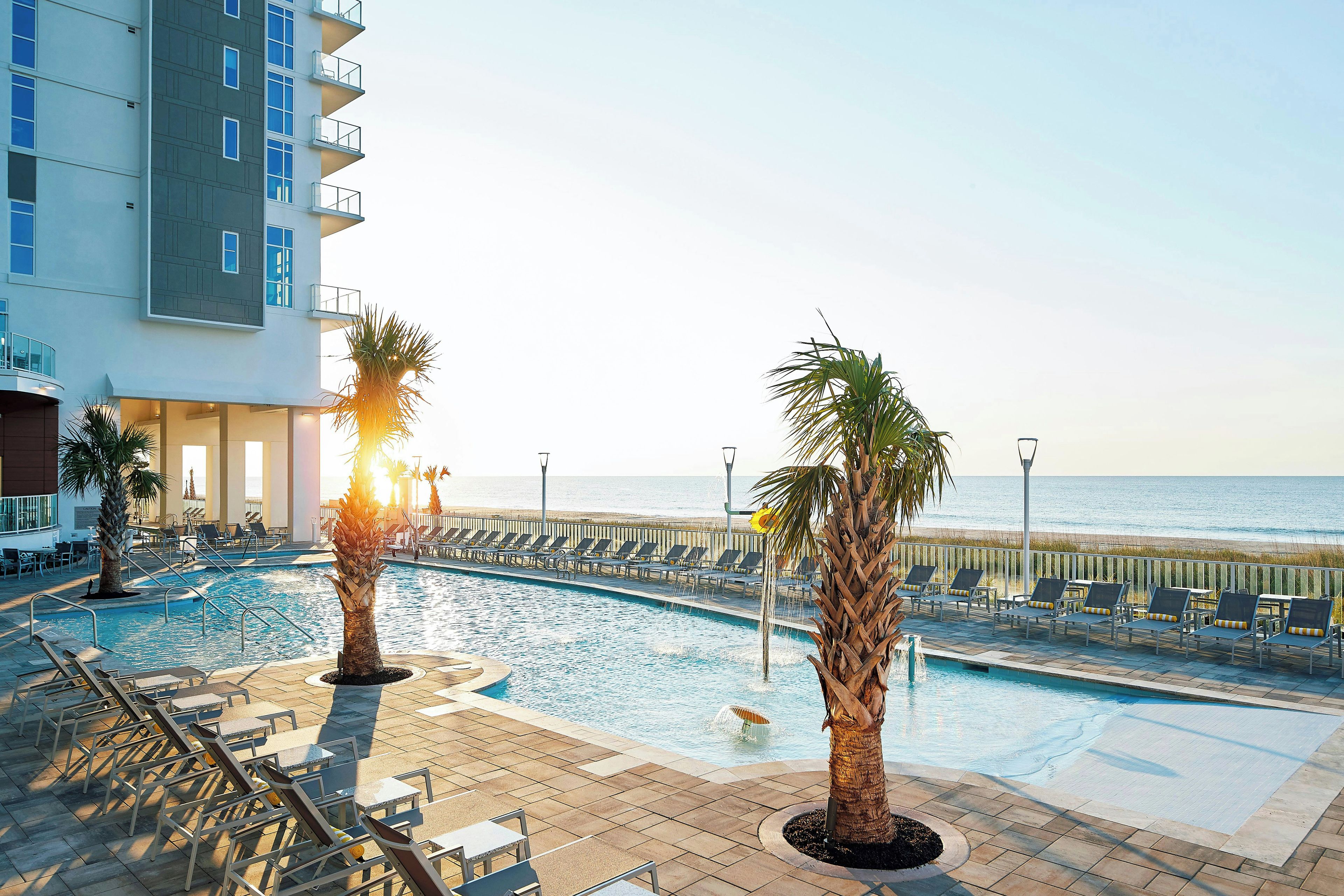 A pool area with lounge chairs and palm trees, overlooking a beach.