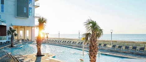 A pool area with lounge chairs and palm trees, overlooking a beach.