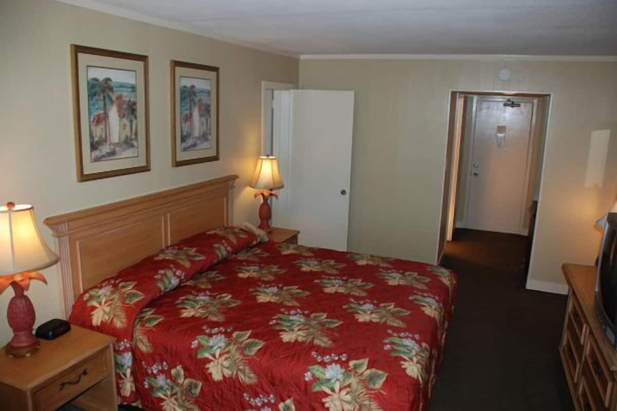 Habitación de hotel con una cama, dos lámparas de noche, una mesita de noche, un aparador y una puerta que da a otra habitación.