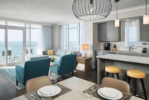 Quarto de hotel moderno com área de jantar, cozinha e grandes janelas com vista para o mar.