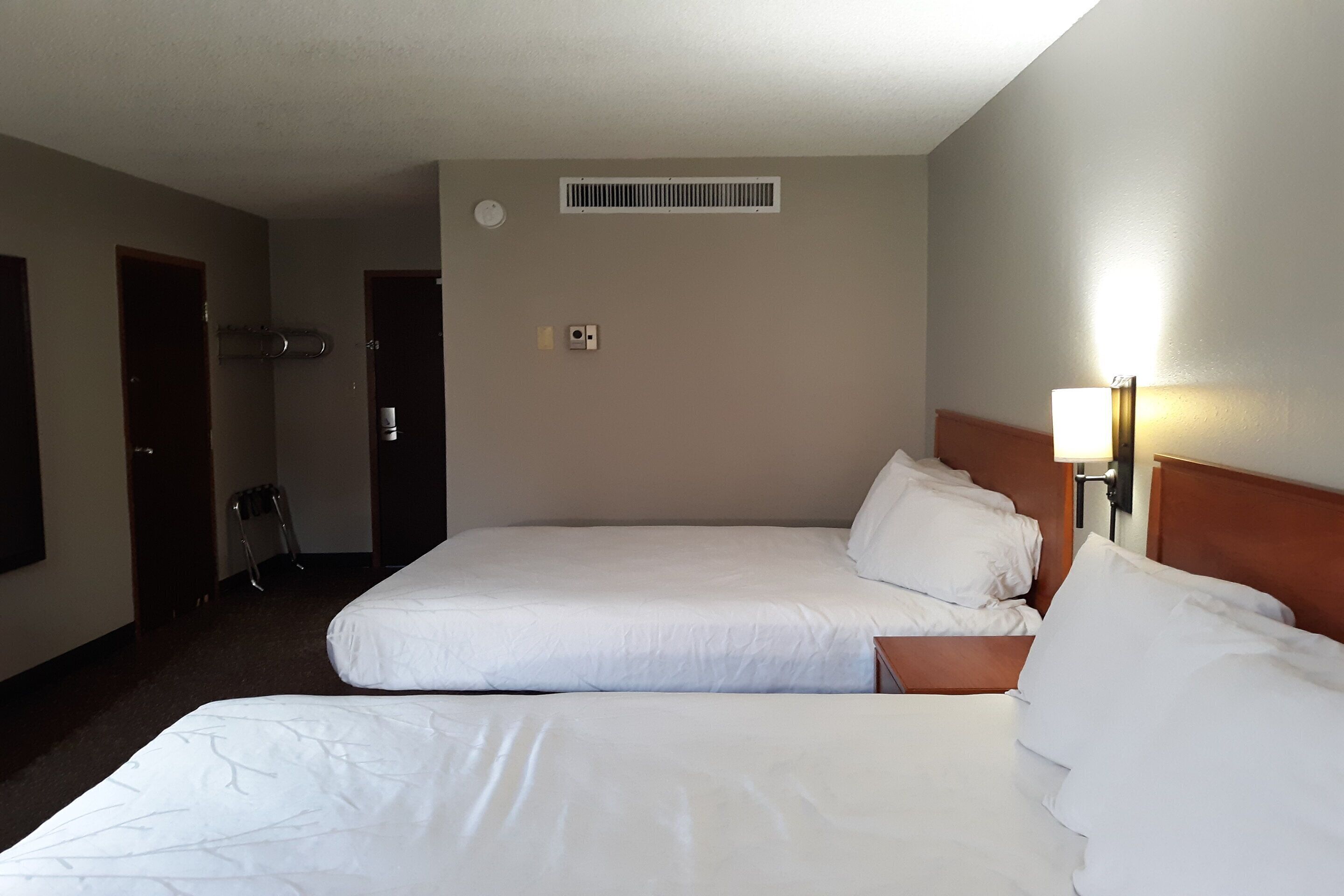 A hotel room with two beds, a wall-mounted heater, a light fixture, and a door.
