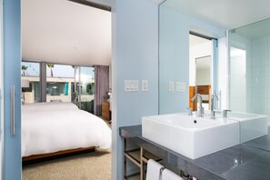 Una habitación de hotel moderna con una cama grande, un baño con un lavamanos y un espejo amplios, y vistas al exterior.