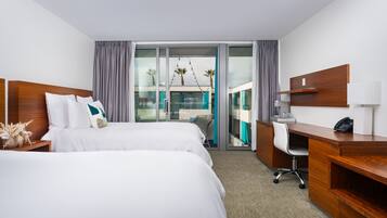 A hotel room with two beds, a desk, and a view of a balcony with palm trees.
