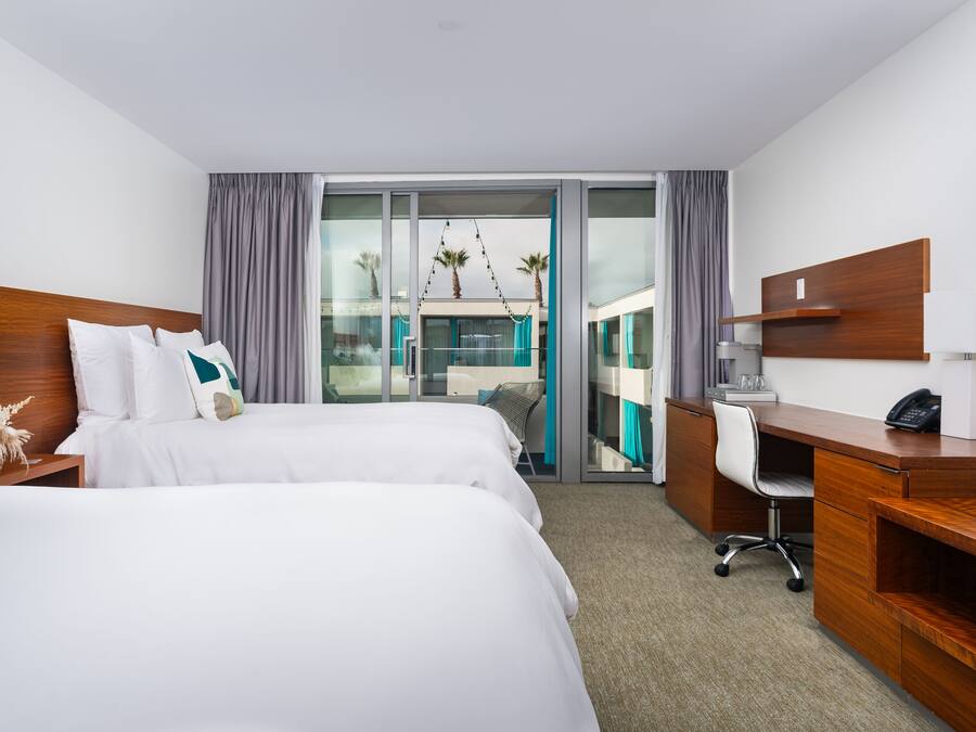 A hotel room with two beds, a desk, and a view of a balcony with palm trees.