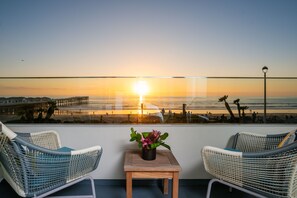 Un balcón con sillones de mimbre y una pequeña mesa con planta en maceta, con vista a una playa y muelle al atardecer.