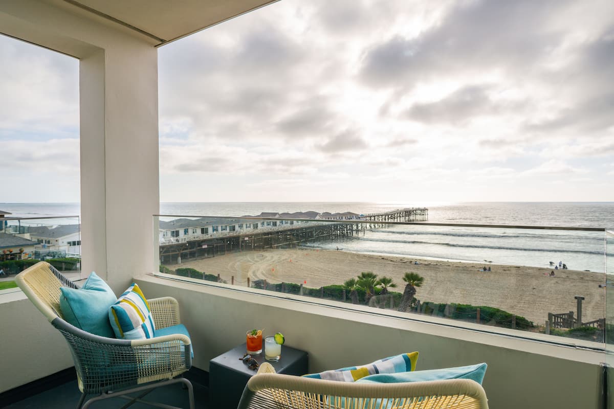 Un balcon avec des fauteuils en osier, une petite table avec une boisson, et une vue sur la plage et la jetée.