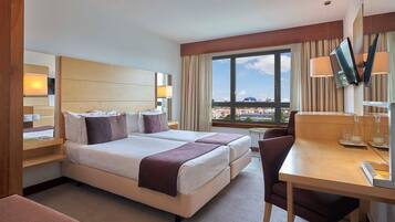 Een hotelkamer met twee bedden, een bureau, een stoel, een televisie en een groot raam met uitzicht over de stad.
