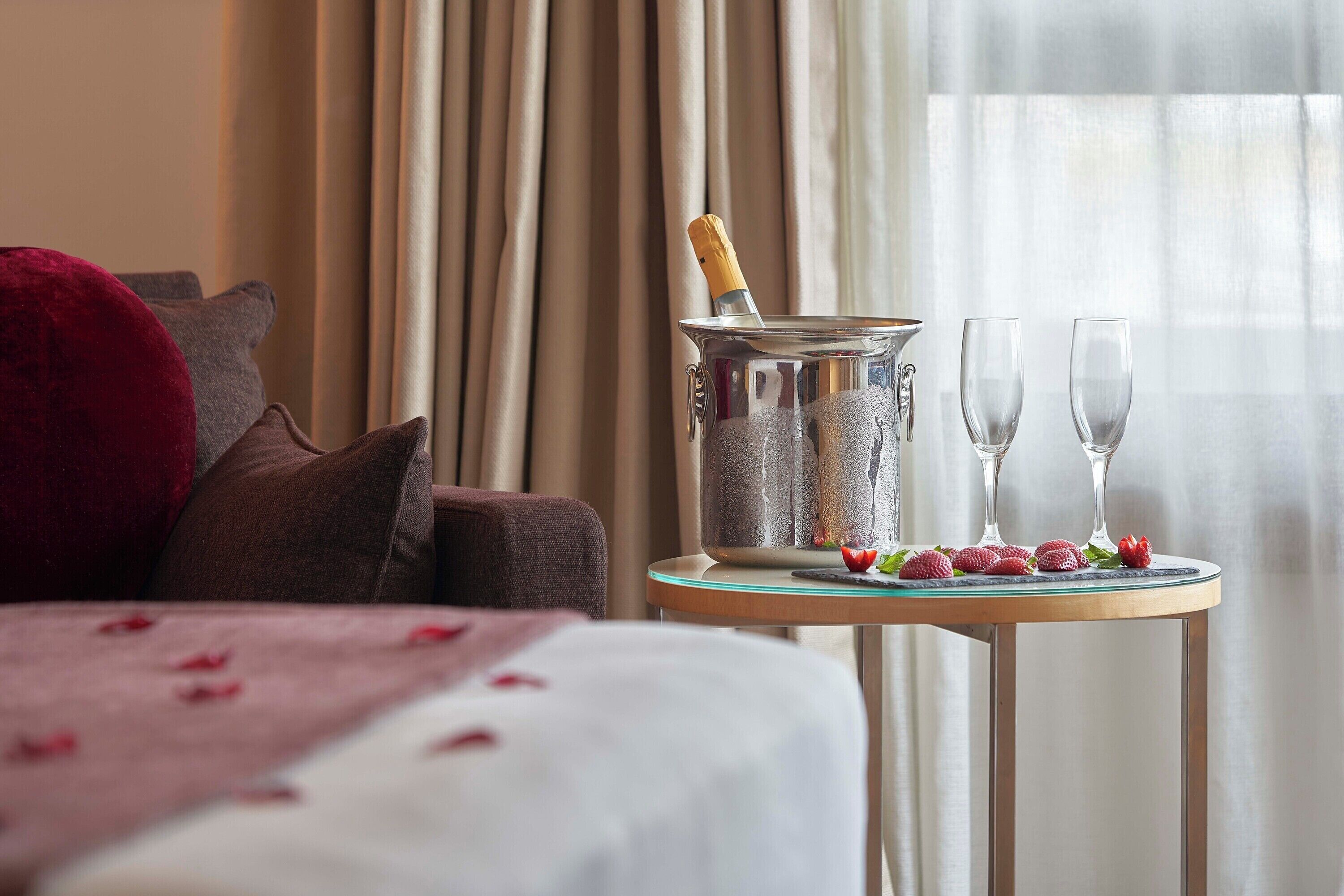 Una habitación de hotel con una cama, una mesita con una botella de champán, dos copas y fresas.