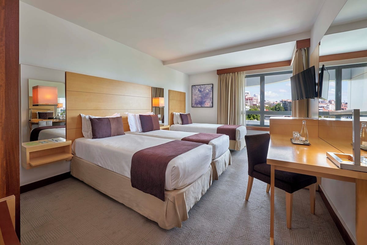 Ein Hotelzimmer mit zwei Betten, einem Schreibtisch, einem Sessel, einem Fernseher und Blick auf die Stadt.