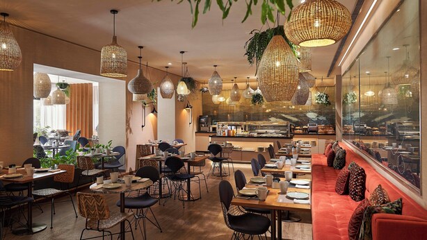 Un interior de restaurante con varias mesas, sillas, lámparas de mimbre colgantes y un banco acolchado rojo a lo largo de la pared.