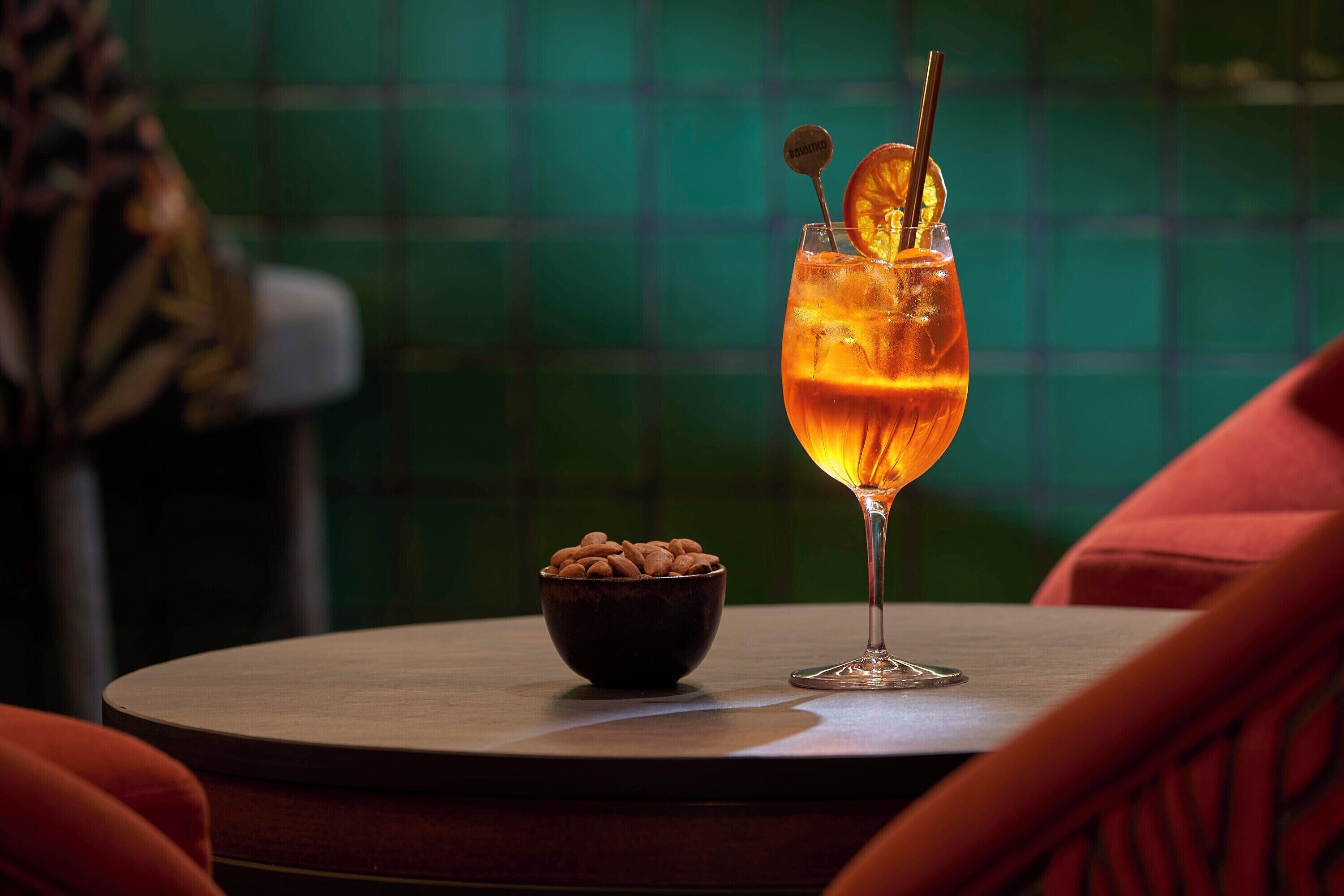 A glass of amber-colored drink with a slice of orange and a straw on a table with a bowl of nuts beside it.