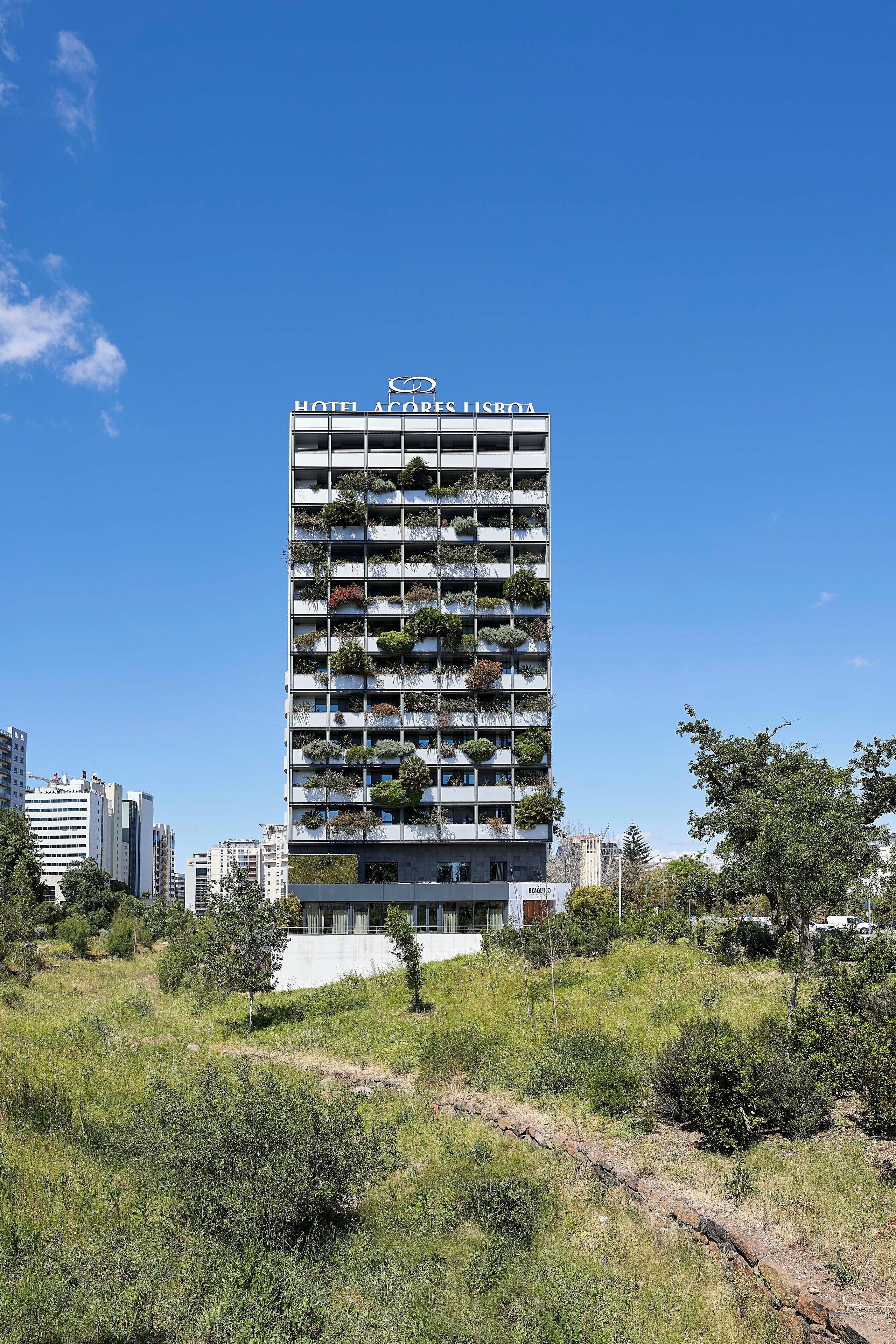 A modern hotel building with balconies, surrounded by greenery and urban buildings.