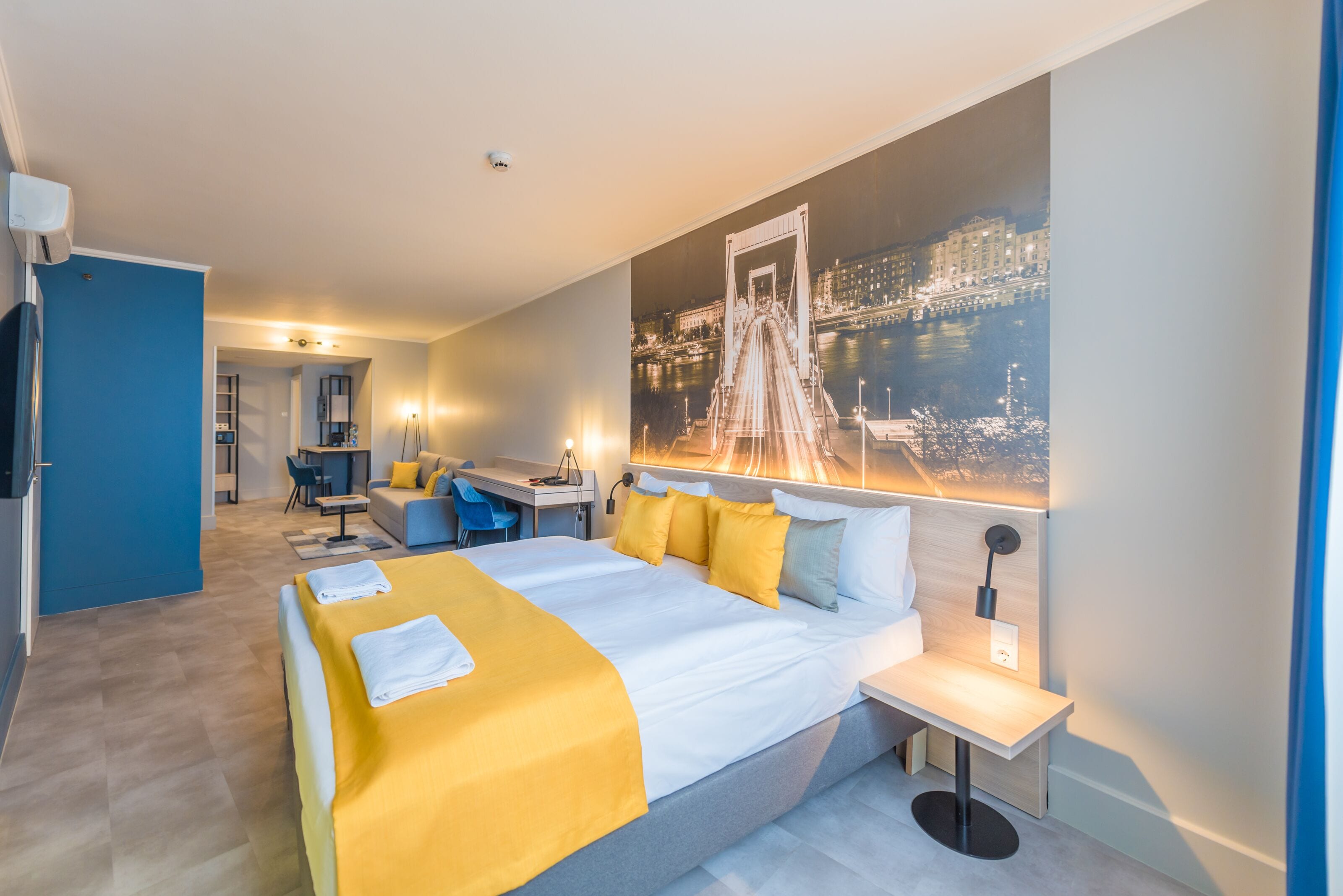 A hotel room with a large bed, a desk, and a chair. There is a painting of a bridge on the wall.