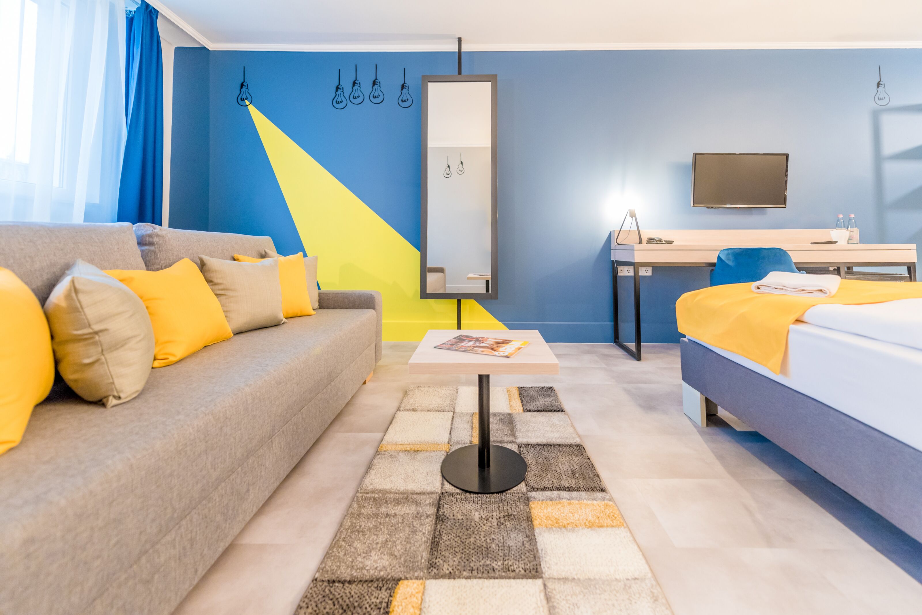 A modern hotel room with a sofa, a bed, a desk, and a large mirror.