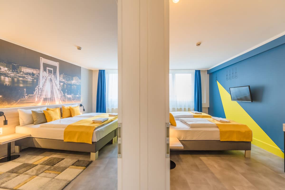 A modern hotel room with two beds, a large window, a cityscape mural, and a flat-screen TV on the wall.