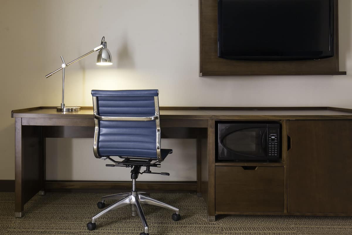 Habitación de hotel con un escritorio de madera, una silla de oficina azul, una lámpara de escritorio, un televisor de pantalla plana y un microondas.