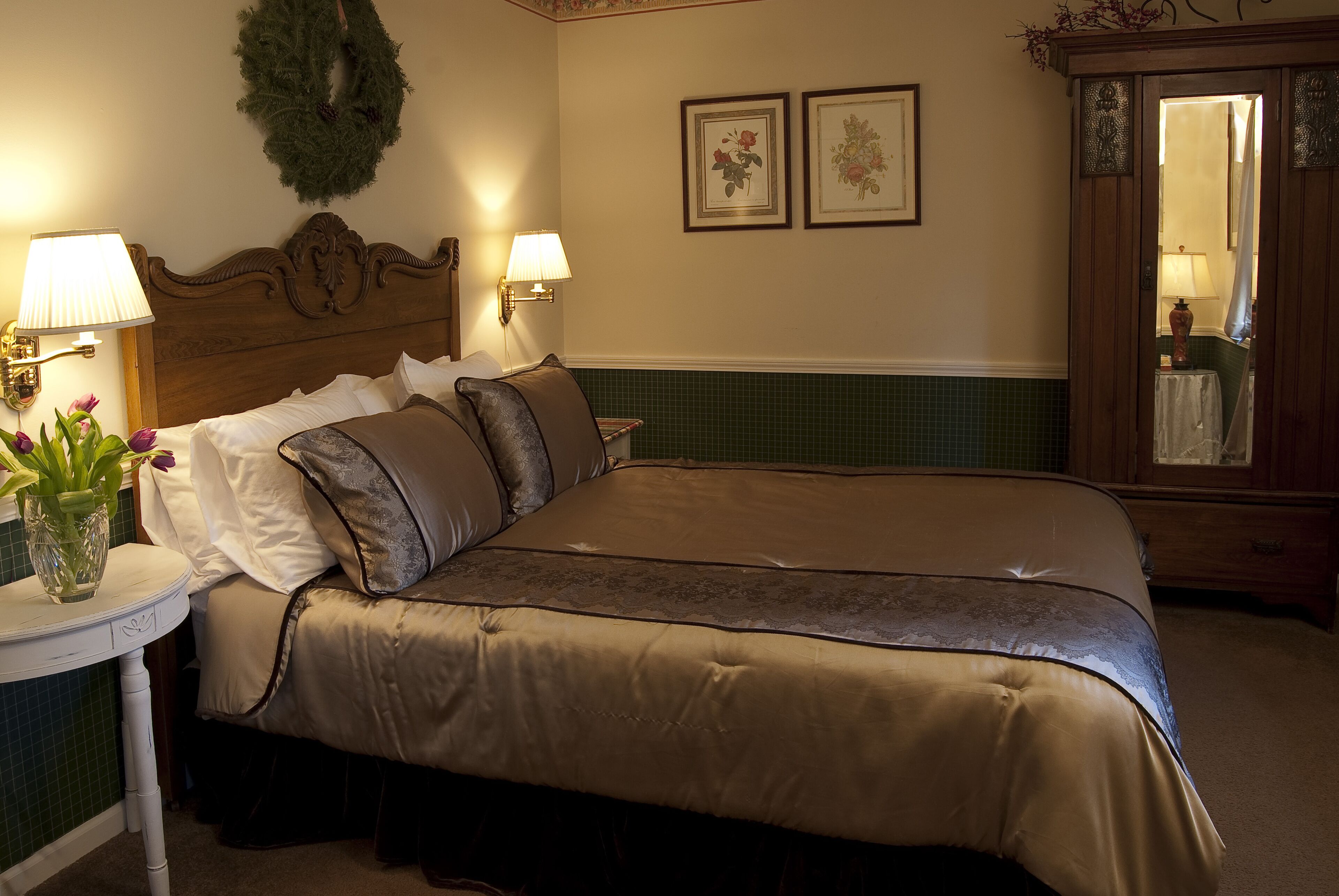 A neatly made bed with a brown bedspread and pillows, a bedside table with a lamp, a vase with flowers, and a mirror cabinet.