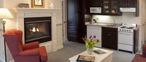 A living room with a fireplace, a red armchair, a white coffee table with a magazine, and a kitchen with dark cabinets and a white stove.