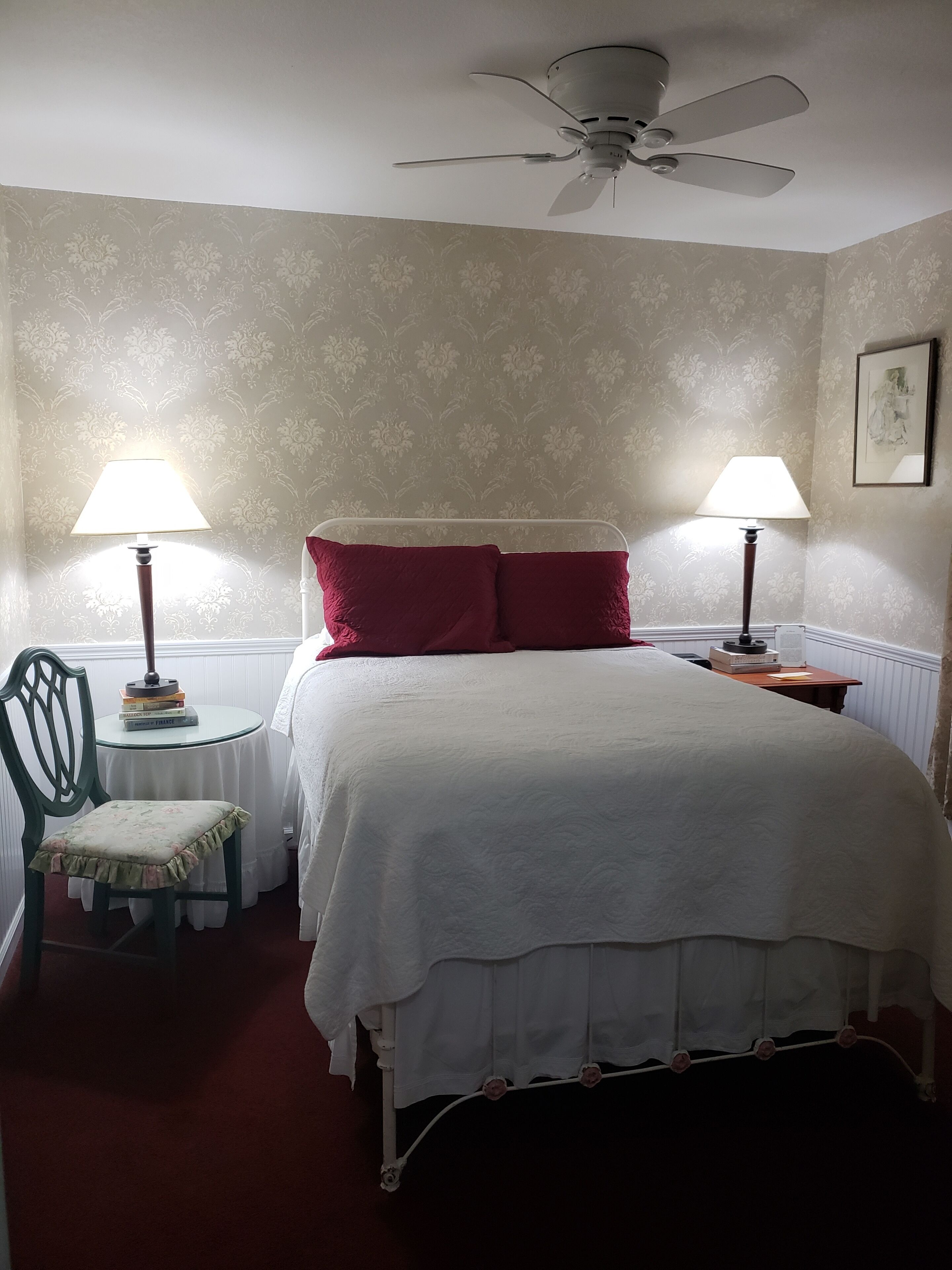 A neatly arranged bedroom with a bed, two bedside lamps, a chair, a small table, and a ceiling fan.