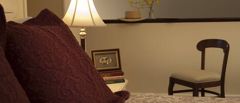 Um quarto com uma cama, luminária de cabeceira, uma cadeira, uma mesinha com livros e porta-retrato, e um vaso com flores em uma prateleira.