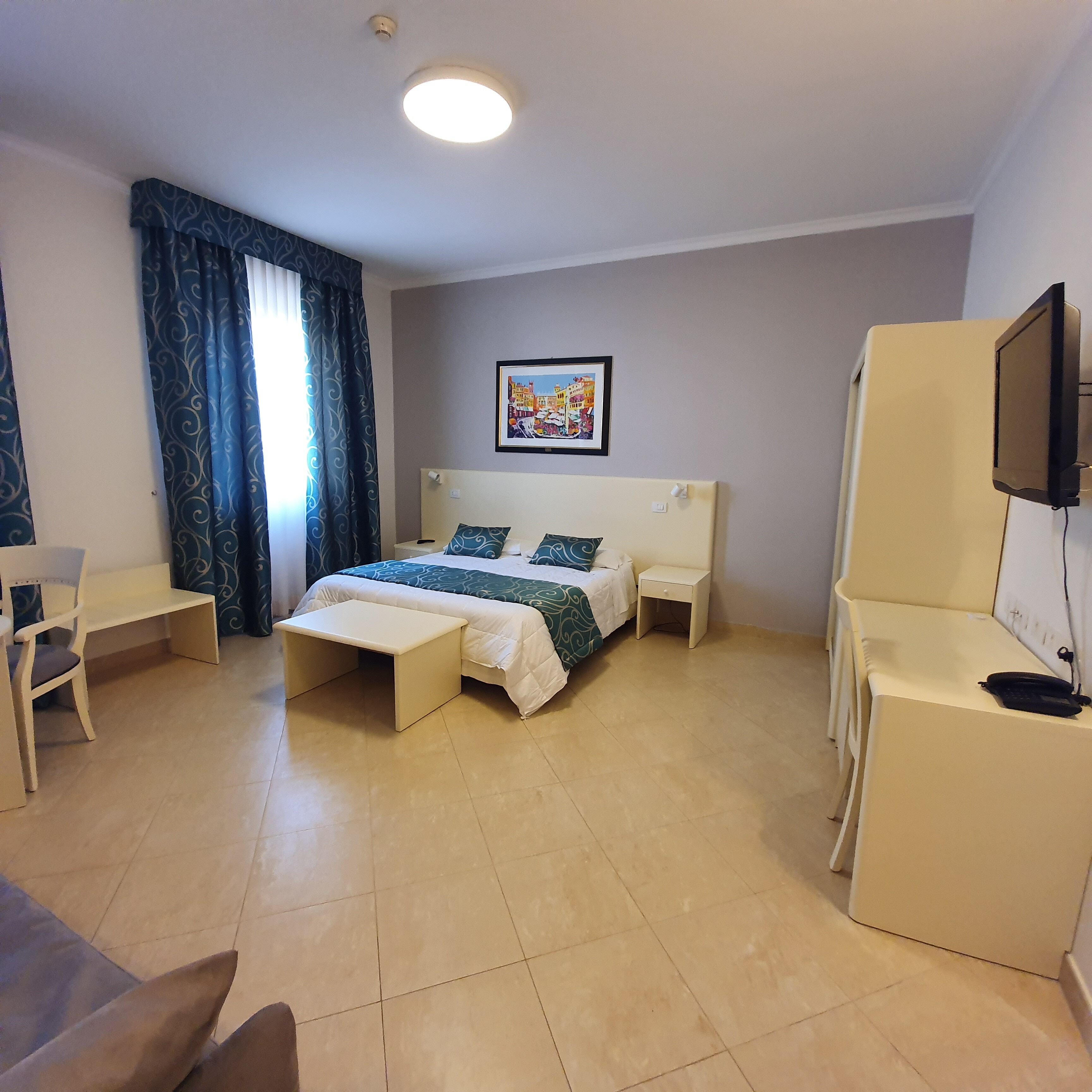 Une chambre d’hôtel comprenant un lit, des tables de chevet, un bureau, une chaise, un téléviseur et une fenêtre avec des rideaux.
