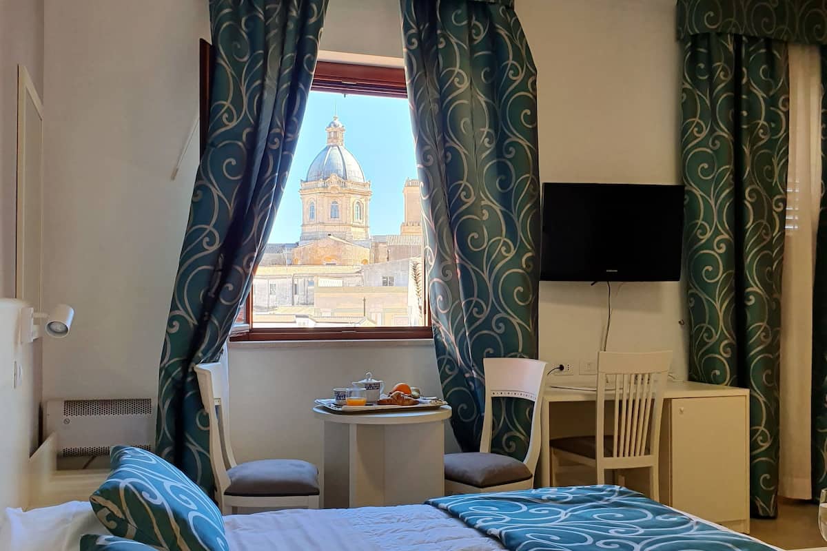 A hotel room with a bed, a desk with a chair, a TV, and a window with curtains.