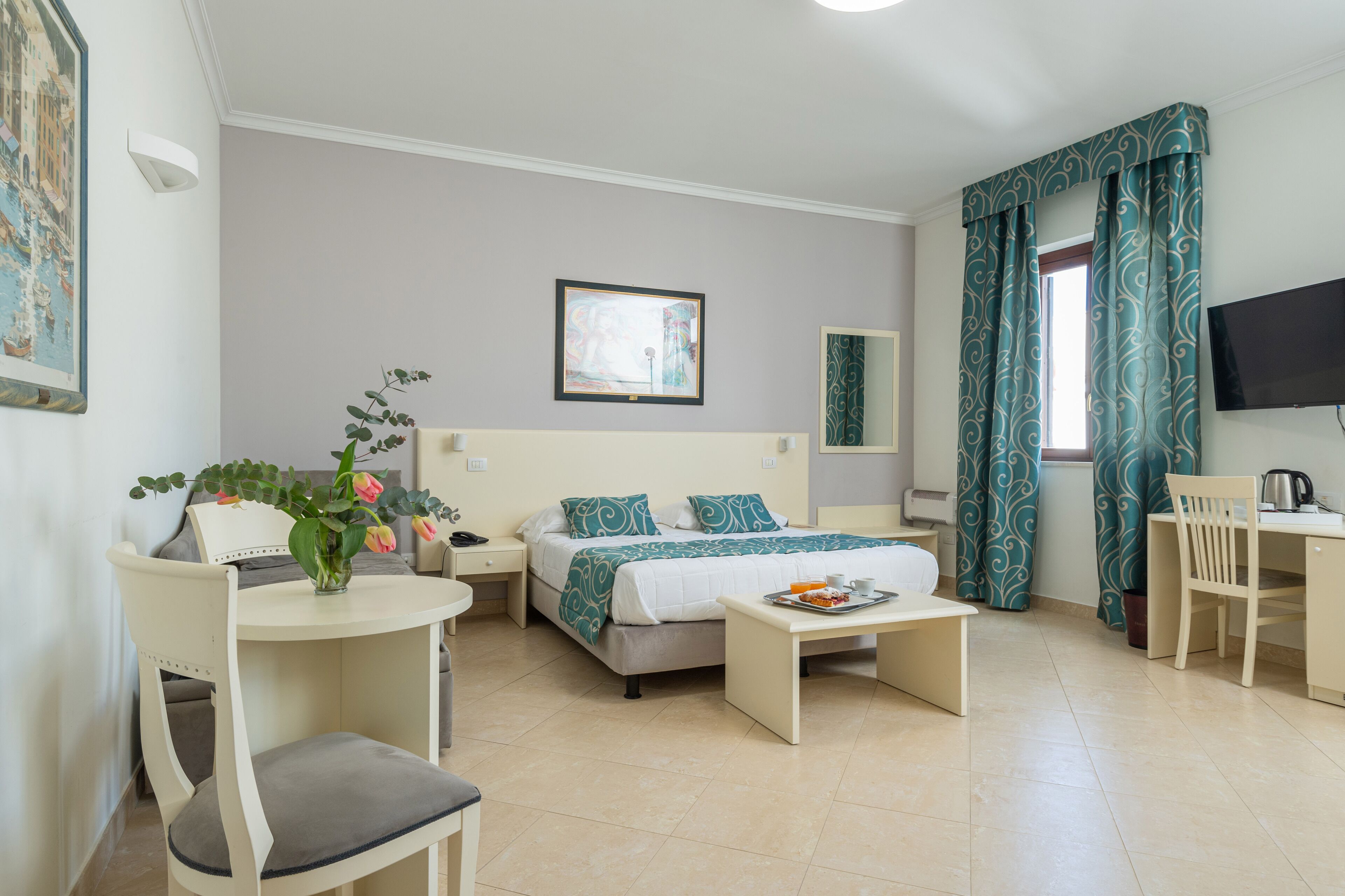 A hotel room with a bed, a desk, a chair, a small table, a TV, and a window with curtains.