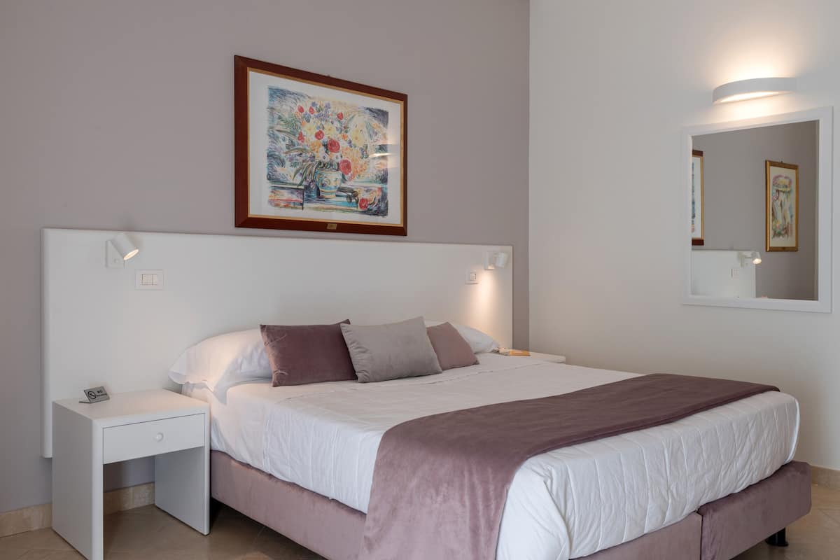 A neatly made bed with a white and brown color scheme, a nightstand with a phone, and a framed painting above the bed.
