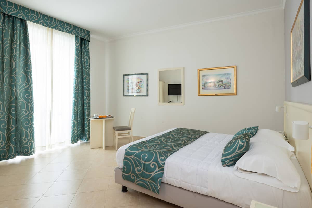 A hotel room with a bed, a chair, a small table, and two framed pictures on the wall.