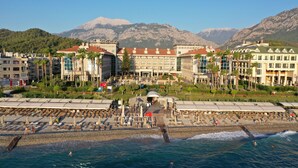 Un hôtel en bord de mer composé de plusieurs bâtiments, une plage avec des transats et des montagnes en arrière-plan.