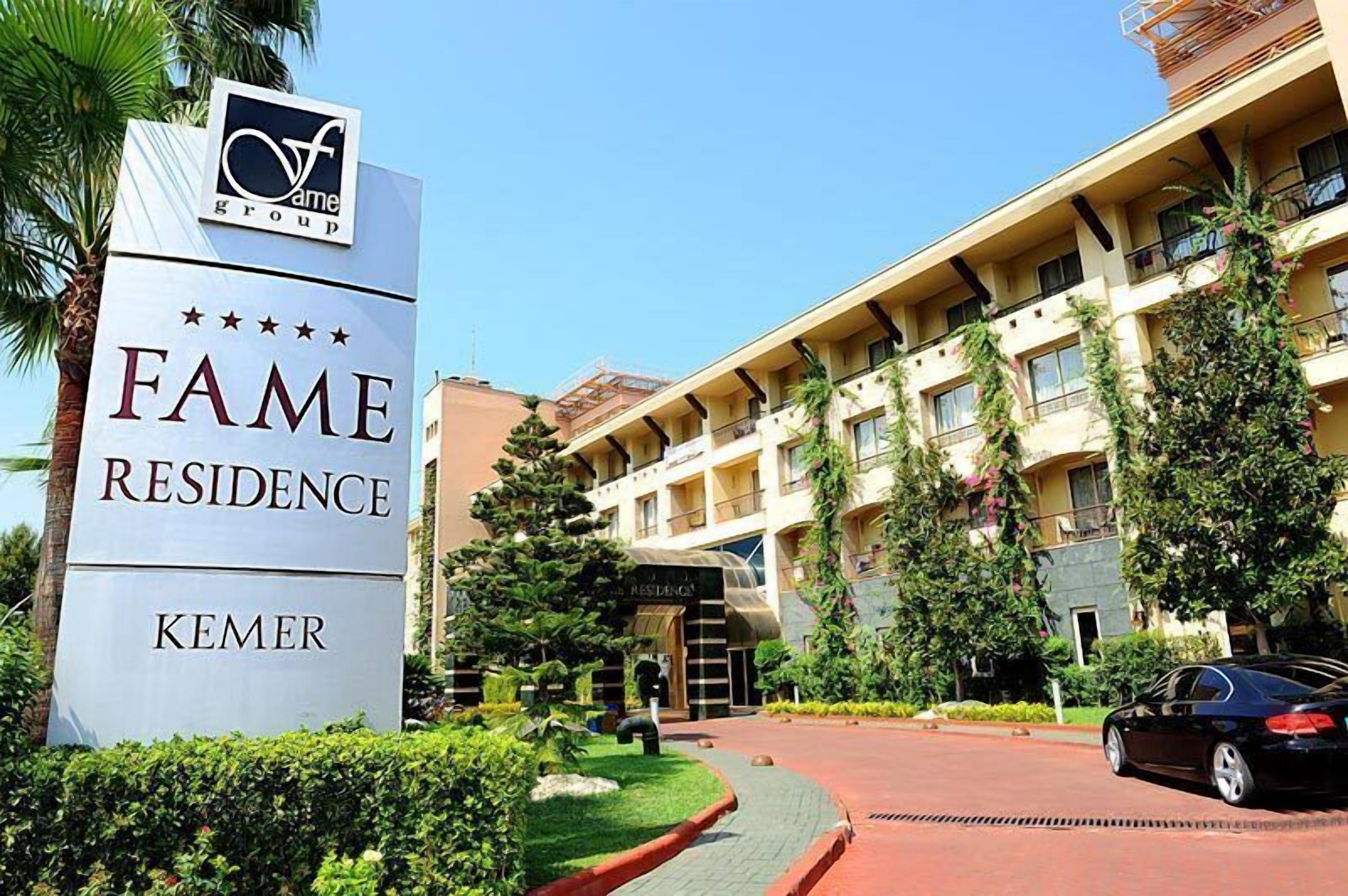 En hotellentrén med en skylt som visar texten ”FAME RESIDENCE KEMER” och en byggnad med flera balkonger och grönska.