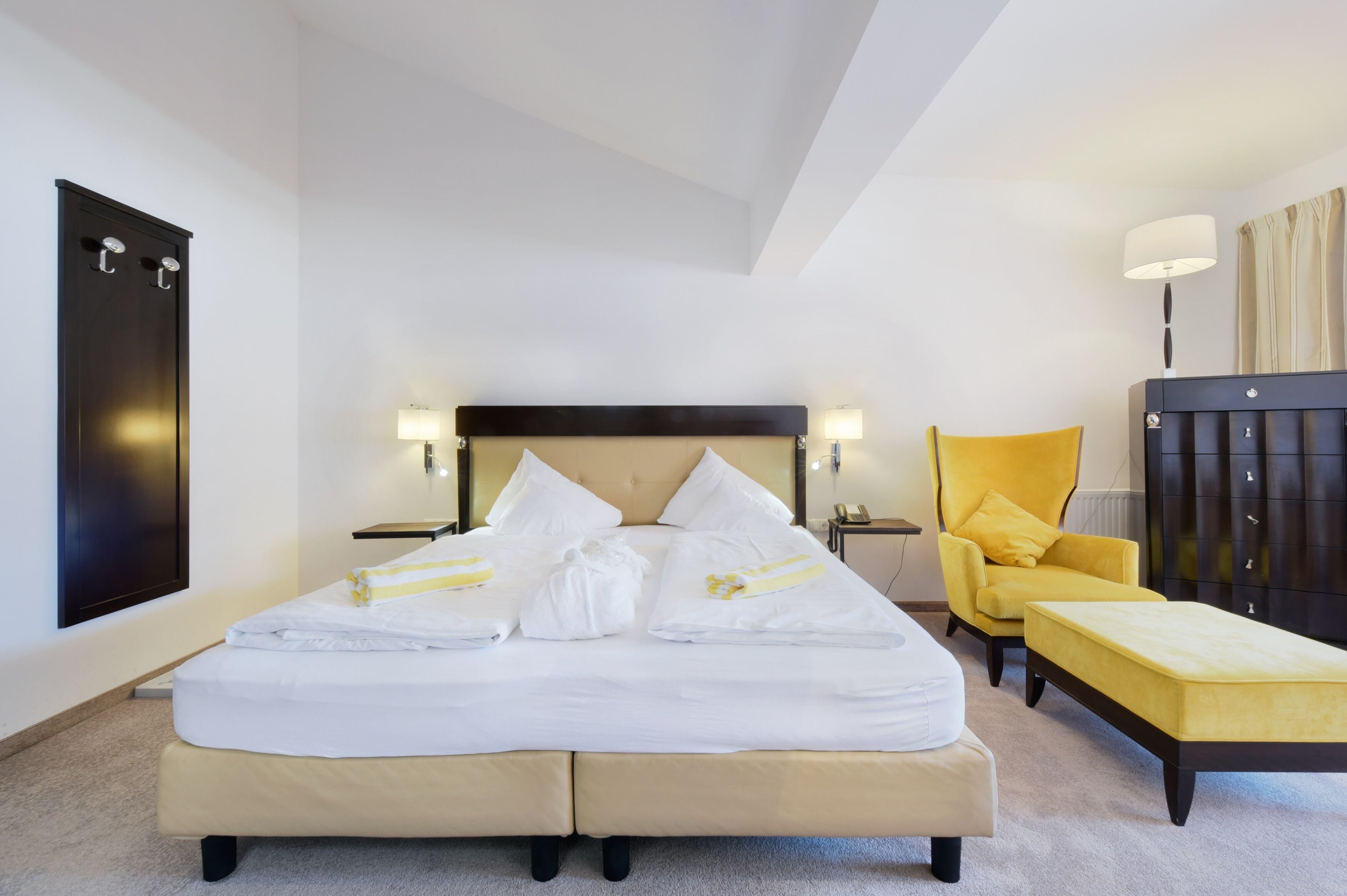 Una cama doble con ropa blanca, una mesita de noche con lámpara, un sillón amarillo y un armario de madera oscura.