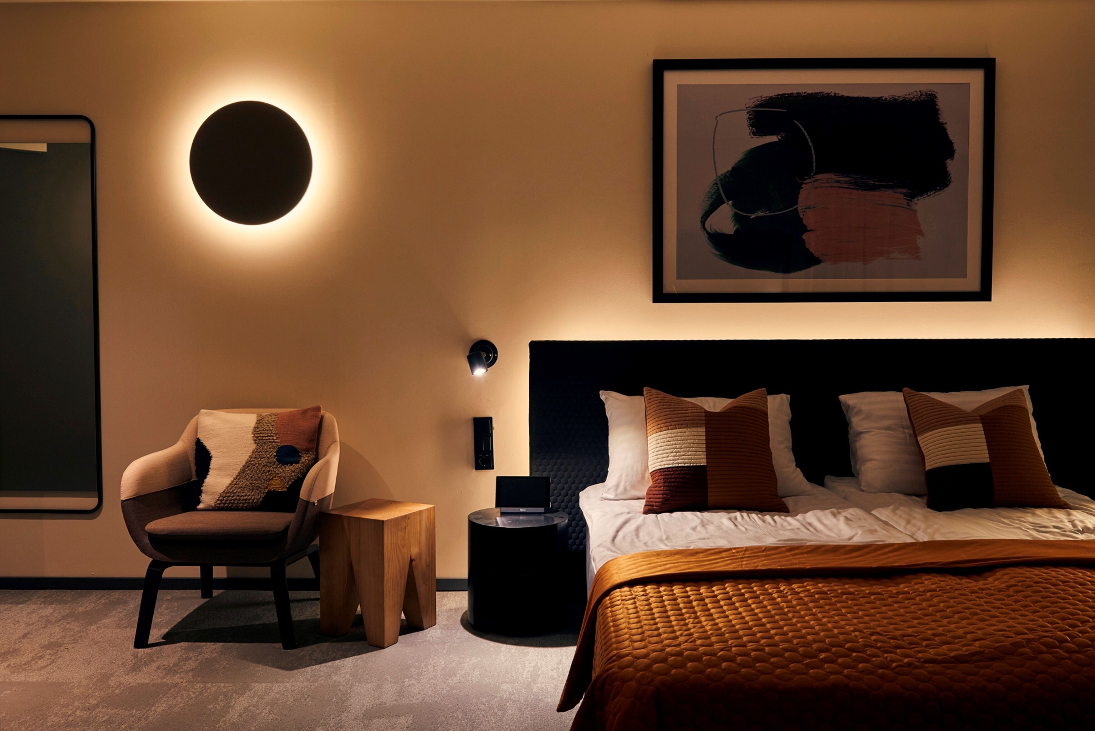 Camera da letto moderna con una lampada a parete rotonda, un quadro astratto incorniciato, un letto con trapunta marrone e una sedia con cuscino decorato.