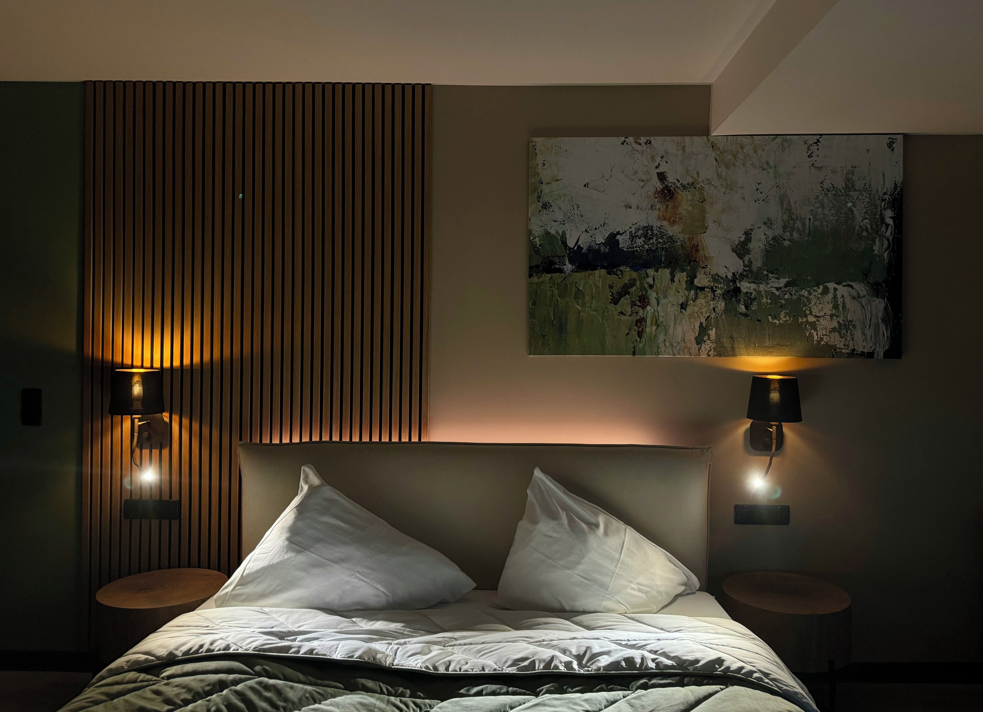 A hotel room with a bed, two bedside lamps, a wooden slat wall, and a painting on the wall.