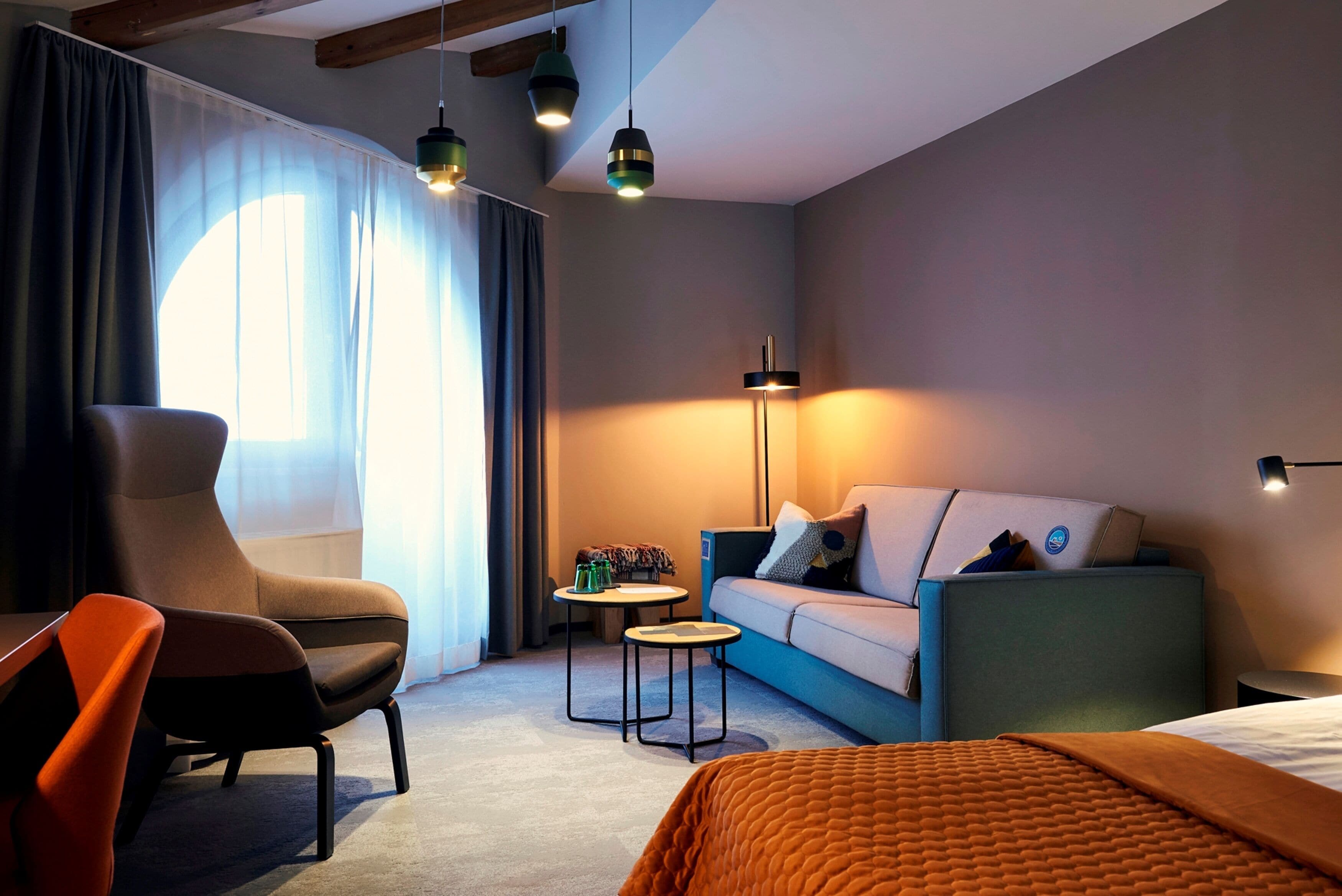 Une chambre d’hôtel moderne, équipée d’un canapé, d’un fauteuil et d’un lit. On y trouve également deux petites tables et une lampe.