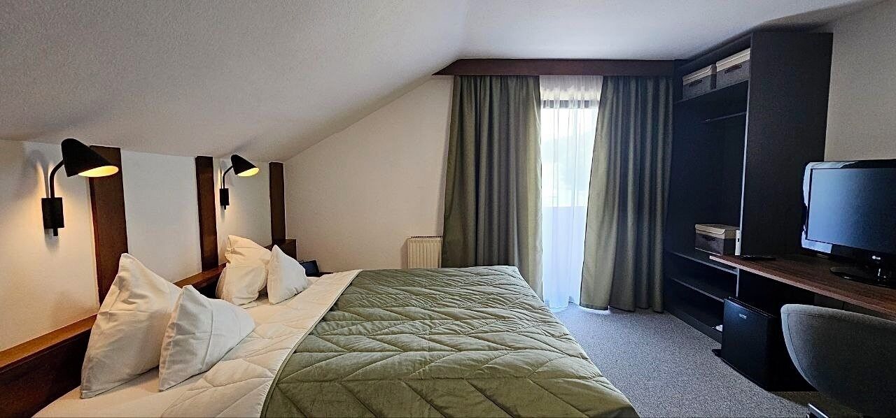 A hotel room with a bed, pillows, a desk with a television, and a window with curtains.