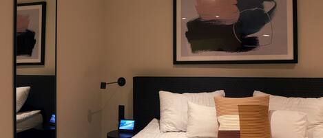 A modern hotel room with a large bed, a bedside table with a lamp, a framed abstract painting above the bed, and a mirror on the wall.