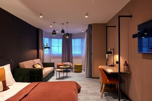 Una habitación de hotel moderna con cama, sofá, escritorio y silla. Hay varias lámparas colgantes y un ventanal con cortinas.